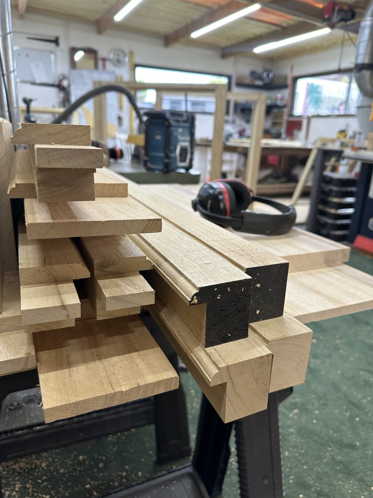 Inside our joinery workshop in Derril — stacked timber stock with profiled mouldings on the bench, ear defenders and a radio in the background